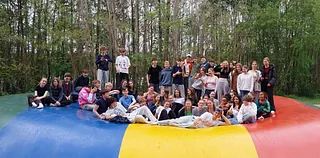 Viele Jugendliche sitzen zusammen auf einem großen bunten Luftkissen draußen im Wald. Alle wirken fröhlich und entspannt. Einige lachen oder machen lustige Zeichen. Das Bild zeigt einen gemeinsamen Ausflug in der Natur.